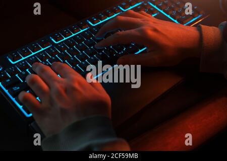 High angle of unrecognizable man in casual clothes typing on black keyboard with comfortable blue backlight while using computer in dark room with dim light at night Stock Photo
