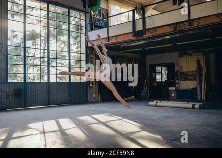 Side view of young slim ballerina in gymnastic suit jumping while practicing dance movements in light spacious studio with large window Stock Photo