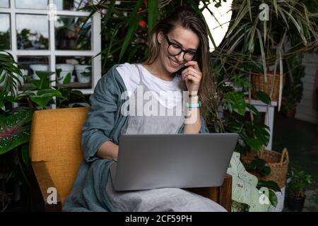 Close up portrait of smiling female gardener in glasses wear linen dress, sitting on chair in green house, using laptop and speaking on web call Stock Photo