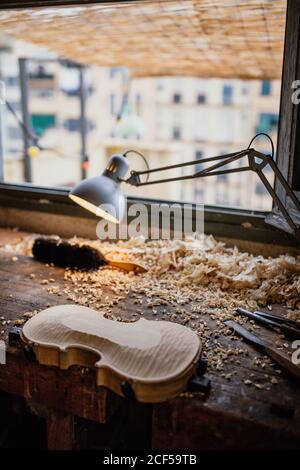 High angle of unfinished violin on wooden table among sawdust beside tools against glowing lamp and window in work studio in daytime Stock Photo