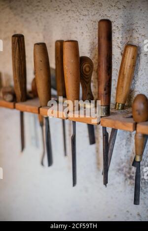 Set of assorted professional carpentry tools with wooden handles placed on shelf hanging on gray plastered wall in workshop Stock Photo