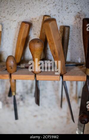 Set of assorted professional carpentry tools with wooden handles placed on shelf hanging on gray plastered wall in workshop Stock Photo