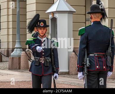 A guardsman of the Royal Guard (His Majesty the King's Guard) gives a hand signal in front of the Royal Palace in Oslo, Norway, while a second guardsman with a rifle stands in the foreground. The palace guard is changed several times a day in a brief ceremony. Stock Photo
