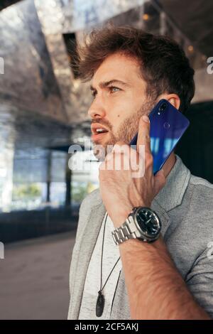 Handsome young male entrepreneur in casual clothing calling on mobile phone and looking away at modern underground car park Stock Photo