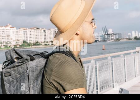 Side view of a man with a nice hat and glasses looking straight ahead carrying a backpack. River and hotels background. Stock Photo