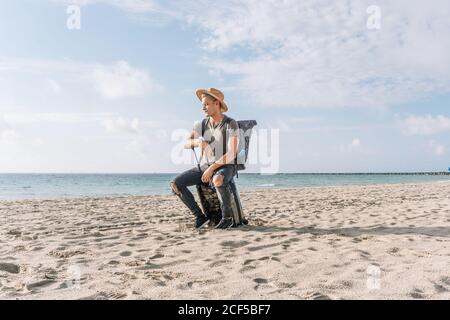 portrait of a lost man sitting on his small suitcase and looking at the sea on the beach, ready for vacation. Stock Photo