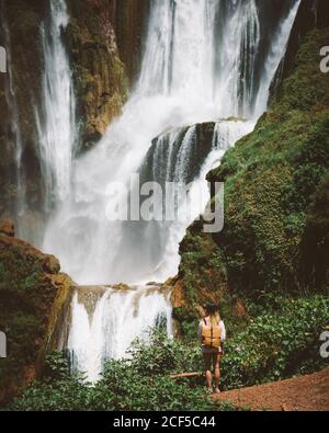Back view of Woman standing in desert terrain with powerful majestic waterfall on green cliffs, Morocco Stock Photo