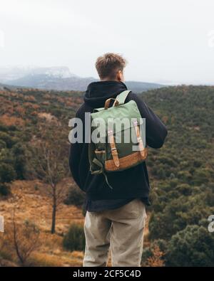 Back view of young male hiker with backpack standing on hill and enjoying beautiful autumn landscape while travelling in mountains Stock Photo