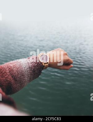 From above crop anonymous female in warm sweater checking time on stylish wristwatch while holding hand over river in cloudy day Stock Photo