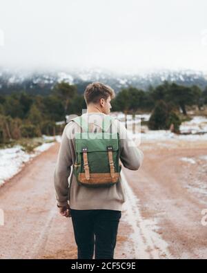 Back view of young male traveler in sweater with backpack walking on dirt rural road in cloudy winter day with green forest and snow on roadsides Stock Photo