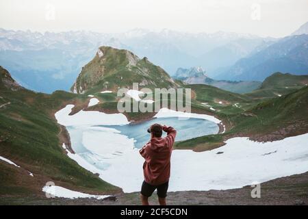 Back view of anonymous male tourist taking photo of mountain lake surrounded by snow and ridge in Pyrenees Stock Photo