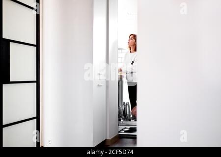 Side view of thoughtful housewife in casual clothes loading dishwasher with tableware while spending weekend at home Stock Photo