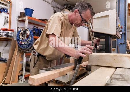 Low angle of attentive middle aged male woodworker in glasses and casual clothes focusing and drilling lumber using special electric machine while working in contemporary workshop Stock Photo