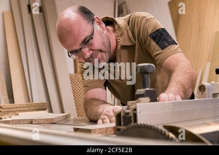 Carpentry master in glasses and casual clothes measuring and sawing timber while crafting detail using electric trimming machine in light contemporary workshop Stock Photo