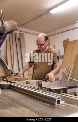 Low angle of focused male woodworker in glasses and casual clothes sawing lumber using special electric trimming machine while working in light modern carpentry studio Stock Photo