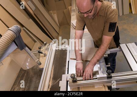 From above crop serious focused carpentry master in glasses and casual clothes measuring and sawing timber while crafting detail using electric trimming machine in light contemporary workshop Stock Photo