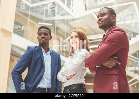 Two handsome young businessmen and lady in classic clothes are looking at camera and smiling, standing with crossed arms near the office building Stock Photo