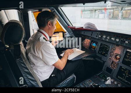 Side view of male captain sitting in cockpit of contemporary aircraft and reading flight document while preparing for departure Stock Photo