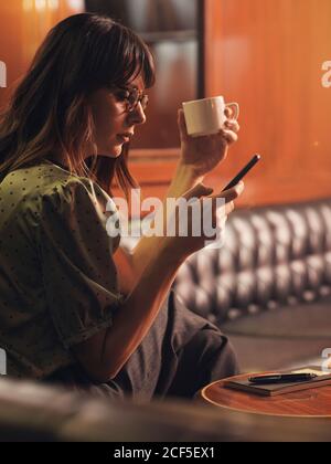 Side view of stylish thoughtful Woman surfing mobile phone and drinking coffee in a comfortable sitting at black leather sofa in cafe Stock Photo