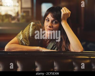 Side view of tired relaxed Woman leaning on back of leather black sofa and looking at camera in cafe Stock Photo