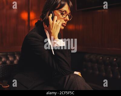 Stylish manager in classical suit talking on mobile phone comfortably sitting on black leather couch in cafe Stock Photo