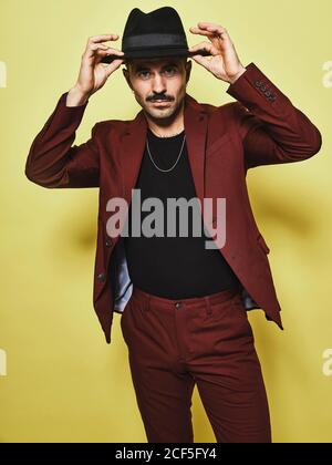 Handsome bearded well dressed male in trendy vinous suit and hat looking at camera against yellow background Stock Photo