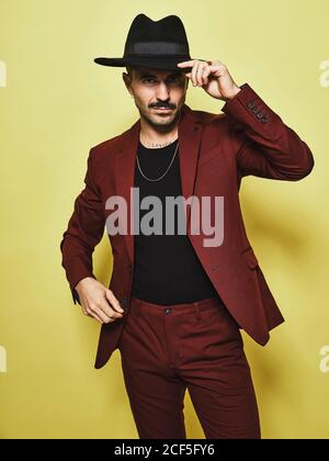 Handsome bearded well dressed male in trendy vinous suit and hat looking at camera against yellow background Stock Photo