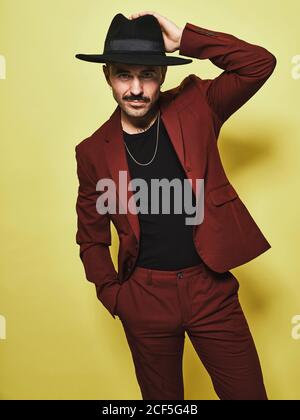 Handsome bearded well dressed male in trendy vinous suit and hat looking at camera against yellow background Stock Photo