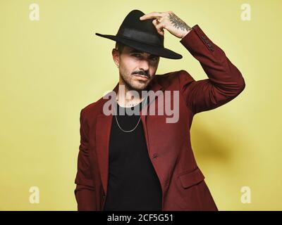 Handsome bearded well dressed male in trendy vinous suit and hat looking at camera against yellow background Stock Photo