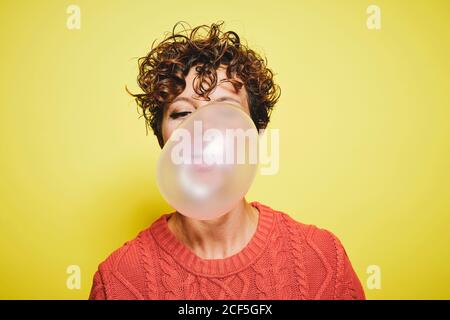 Young curly haired female in orange sweater blowing bubble gum while standing against yellow background Stock Photo