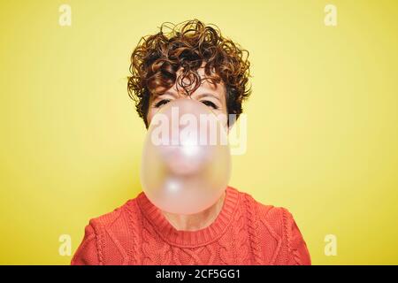 Young curly haired female in orange sweater blowing bubble gum looking to the camera while standing against yellow background Stock Photo