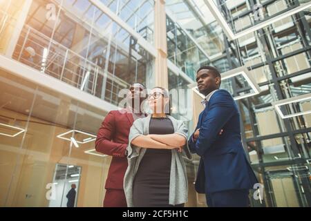 Two handsome young businessmen and lady in classic clothes are looking at camera and smiling, standing with crossed arms near the office building Stock Photo