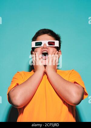 Shocked surprised preteen boy with mouth opened in special glasses with stereoscopic blue and red lens looking away and enjoying 3d graphic video against turquoise background in modern studio Stock Photo