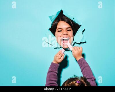 Cheerful little girl standing behind blue paper sheet and looking through hole eating marshmallow from hands of infant kid Stock Photo