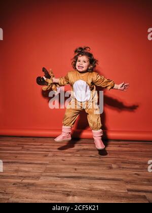 Energetic happy little girl in cute deer costume spreading arms while jumping looking up against red wall in studio Stock Photo