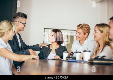 Welcome on board Two handsome men shaking hands with smile while sitting on the couch at office with their coworkers Stock Photo