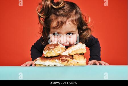 Closeup portrait of cheerful little girl enjoying sweet eclairs with chocolate while looking away sitting on the table against red background Stock Photo