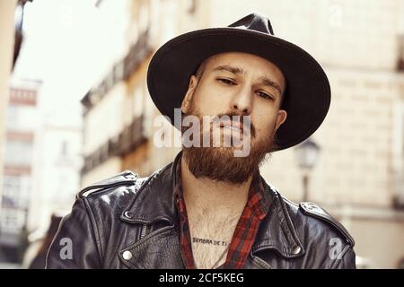 Young bearded handsome man in black hat and leather jacket thoughtfully looking at camera on background of city street Stock Photo