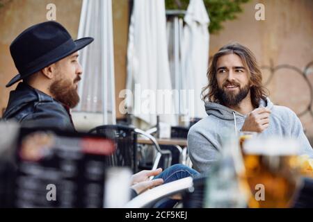 Handsome man in black hat sitting and enjoying conversation with friend wearing gray hoodie in outside cafe Stock Photo