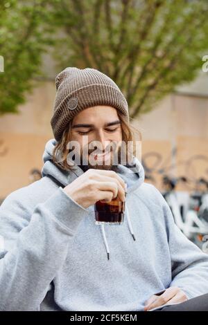 Young bearded handsome man in grey hoodie sitting holding glass of drink and laughing Stock Photo