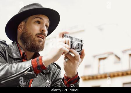 Young bearded handsome man in black hat and leather jacket taking photo in street Stock Photo