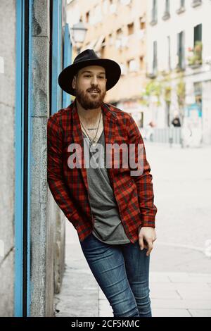 Young bearded handsome man in hat and checkered shirt smiling and looking along standing leaning on wall Stock Photo