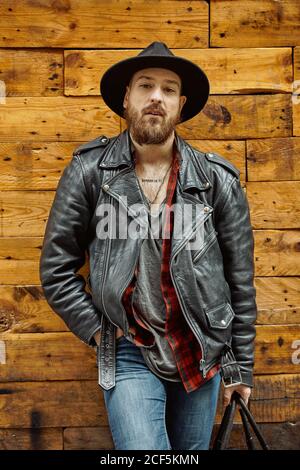 Young bearded handsome man in hat checkered and leather jacket shirt smiling and looking along standing leaning on wooden wall Stock Photo