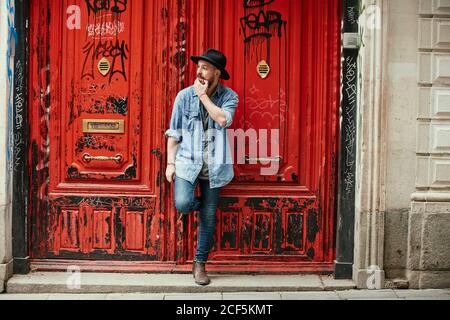 Young bearded handsome man in hat and denim shirt looking along standing leaning foot into red painted doors Stock Photo