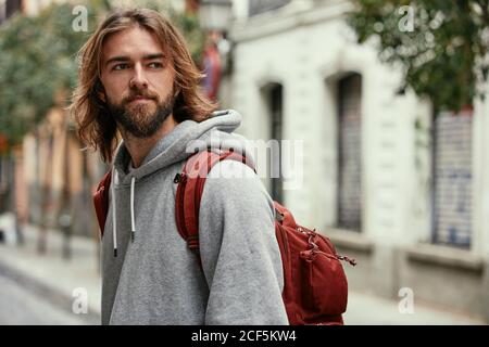 Young bearded handsome man in grey hoodie with backpack seriously walking outside Stock Photo