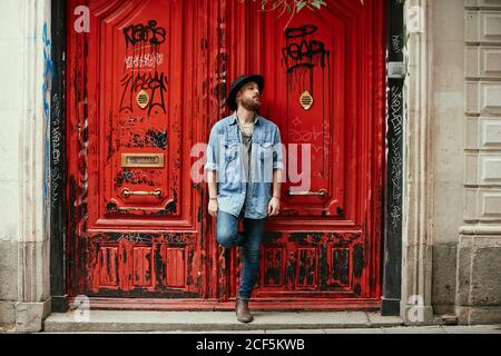 Young bearded handsome man in hat and denim shirt looking along standing leaning foot into red painted doors Stock Photo