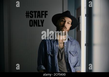 Young bearded handsome man in hat and denim shirt smiling and looking at camera in dark room Stock Photo
