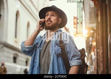 Young bearded handsome man in black hat and denim jacket merrily talking on mobile phone in street Stock Photo