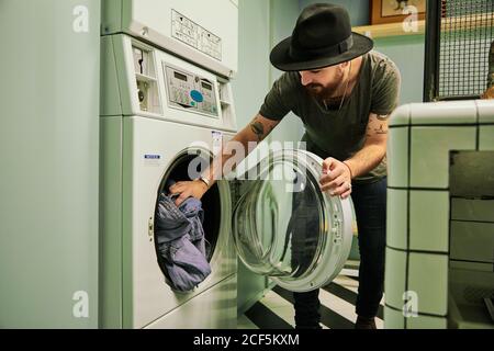 Young bearded handsome man in hat putting things in washing machine in laundry room Stock Photo
