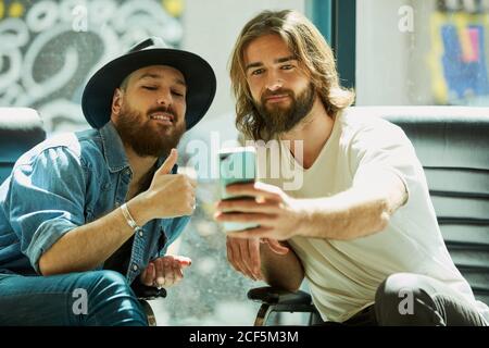 Handsome man in black hat sitting and enjoying process of taking selfie on mobile phone with friend in cafe Stock Photo